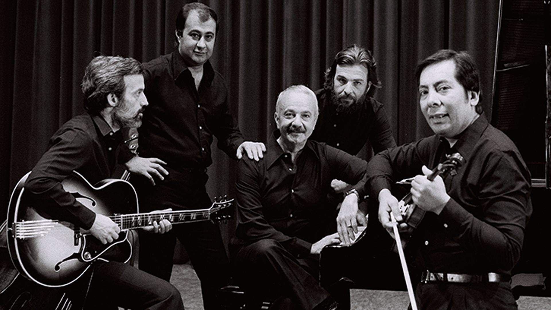 Cinco hombres con camisas negras posan juntos; uno sostiene una guitarra, otro un violín.