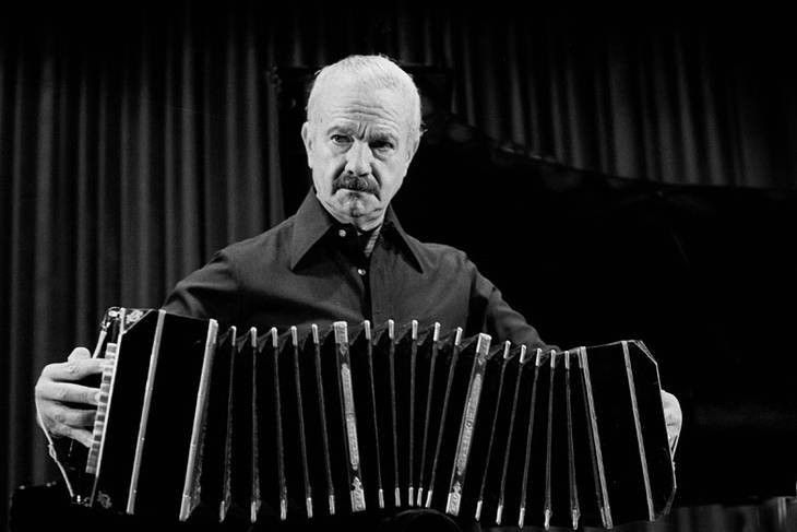 Hombre con expresión seria, sosteniendo un bandoneón frente a un piano. Foto en blanco y negro.