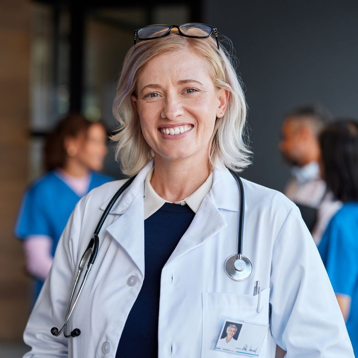 Smiling doctor in a white coat with a stethoscope, standing in a hospital corridor with colleagues blurred behind.