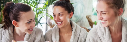 Three women smiling and talking together in white robes near a leafy plant