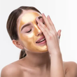 Woman applying gold face mask to her face with eyes closed on a white background