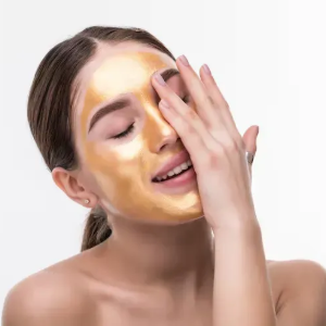 Woman applying gold face mask to her face with eyes closed on a white background