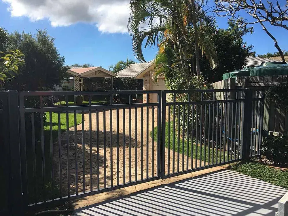 Black metal gate in front of a driveway leading to houses, under a blue sky with green trees — Sunshine Gates in Caloundra, QLD