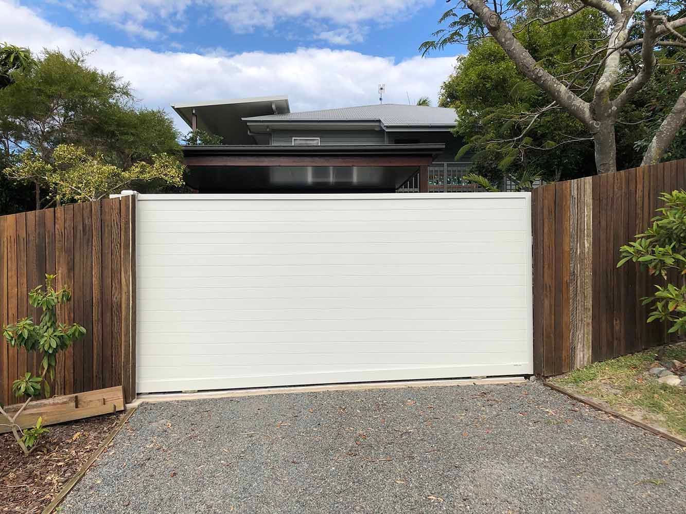 A White Sliding Gate is Leading to a House Behind a Wooden Fence — Sunshine Gates in Kunda Park, QLD