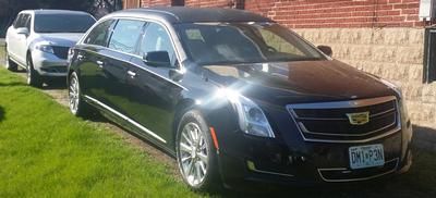 Black Cadillac SUV parked on grass beside a brick wall, with a white car behind it.