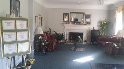 Bright living room with fireplace, blue carpet, framed certificates, and plants by the windows.