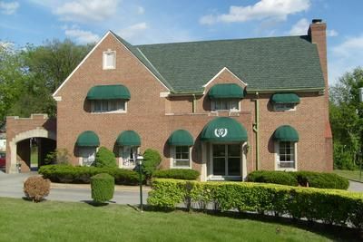 Brick house with green awnings, manicured hedges, and a covered front entrance under a blue sky