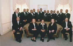 Group of formally dressed people posing for a portrait in a bright room with draped windows.