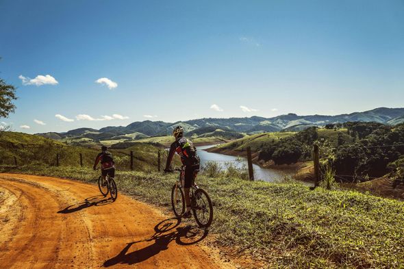 Two cyclists on a dirt road overlooking lush green hills and a distant river under a blue sky