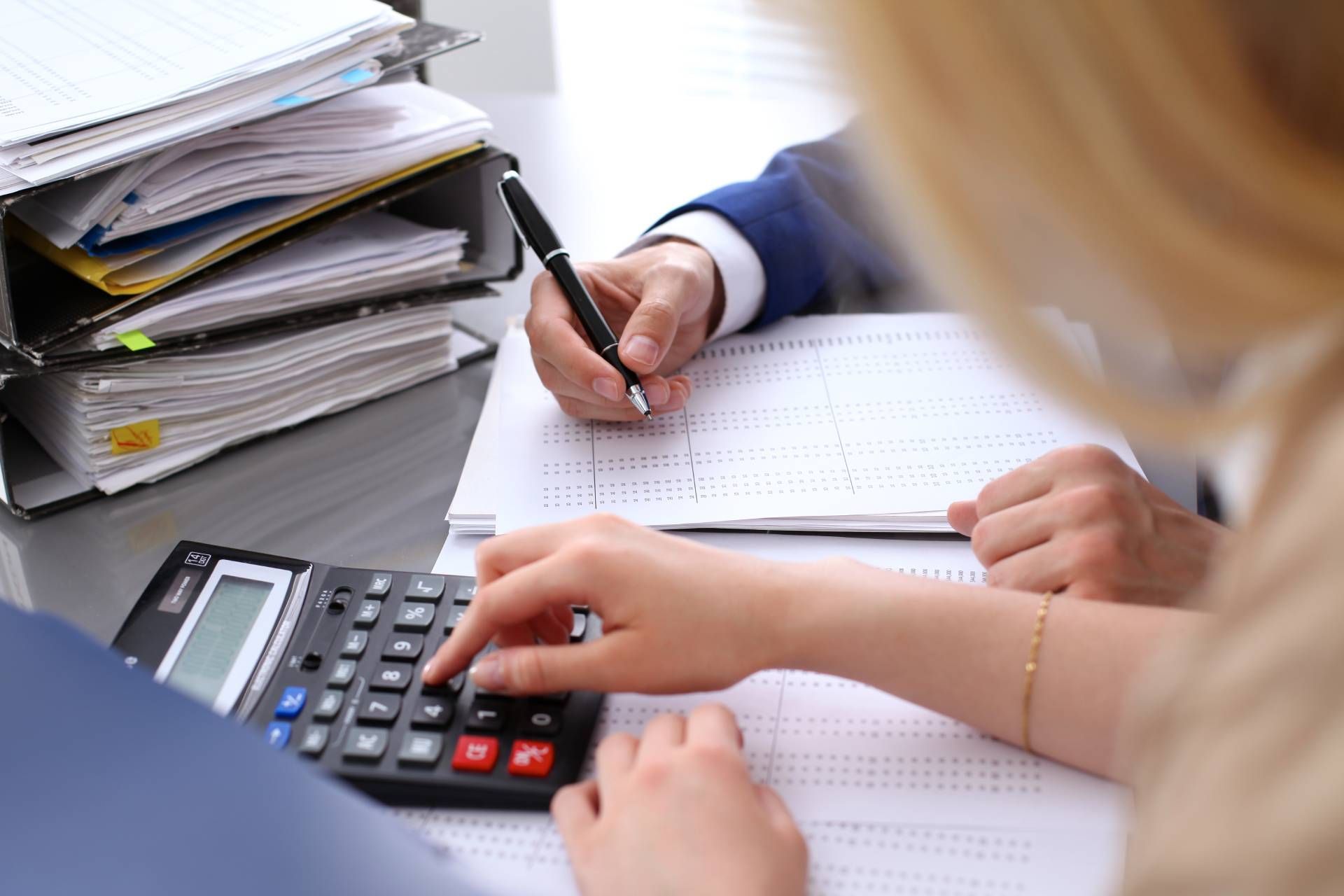 Hands calculating with a calculator and writing on paper; files stacked nearby.