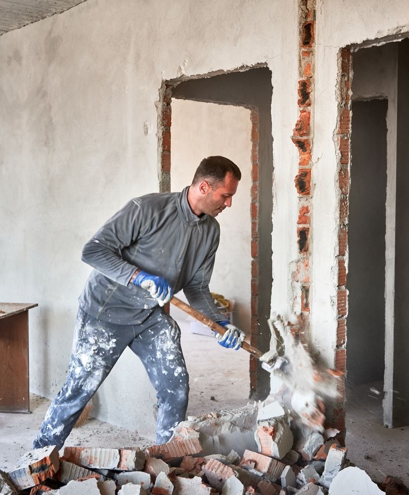 Man demolishing a brick wall with a tool inside a room, surrounded by rubble and dust.