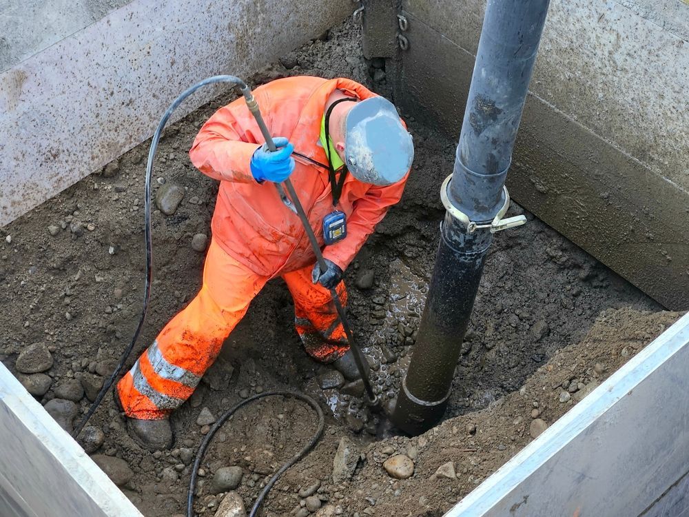 Worker in orange jumpsuit uses a tool near a black pipe in a concrete enclosure.