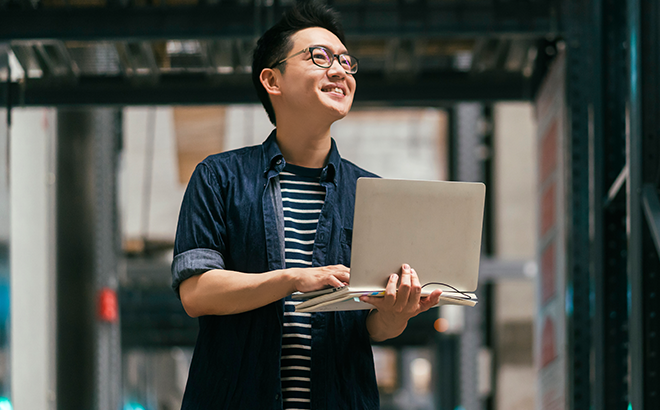 A man is holding a laptop computer in a warehouse.