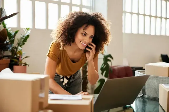 A woman is talking on a cell phone while using a laptop computer.