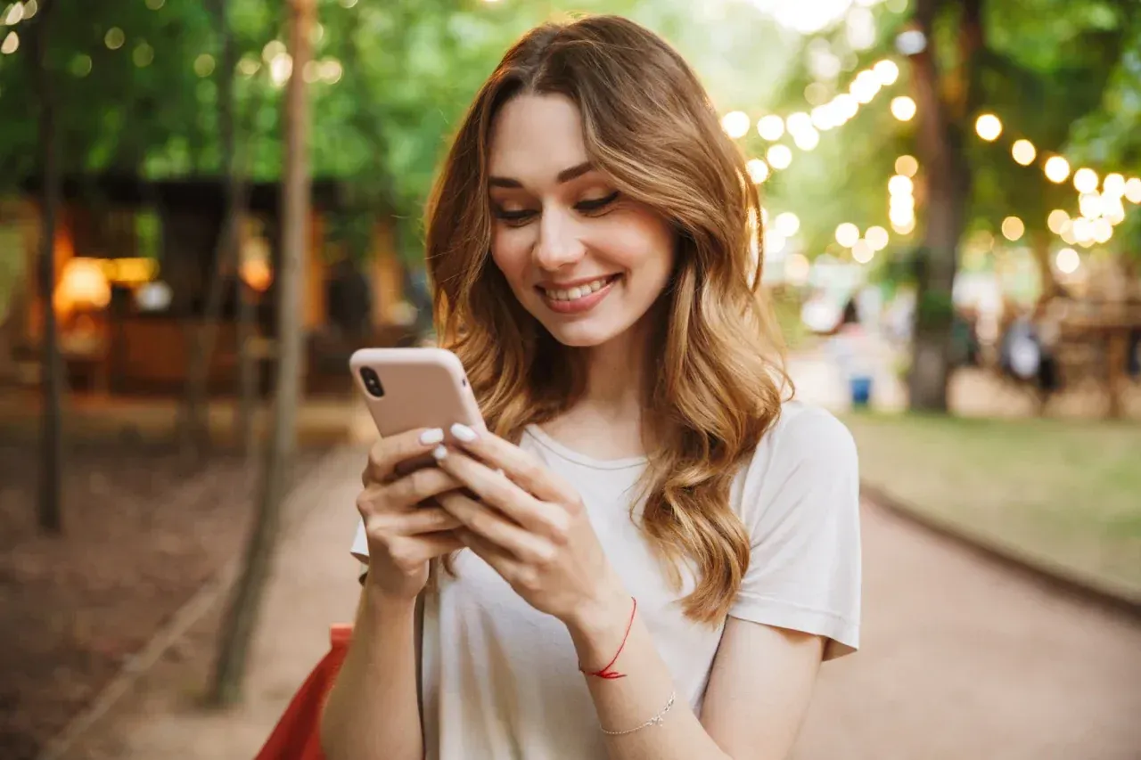 A woman is smiling while looking at her cell phone in a park.