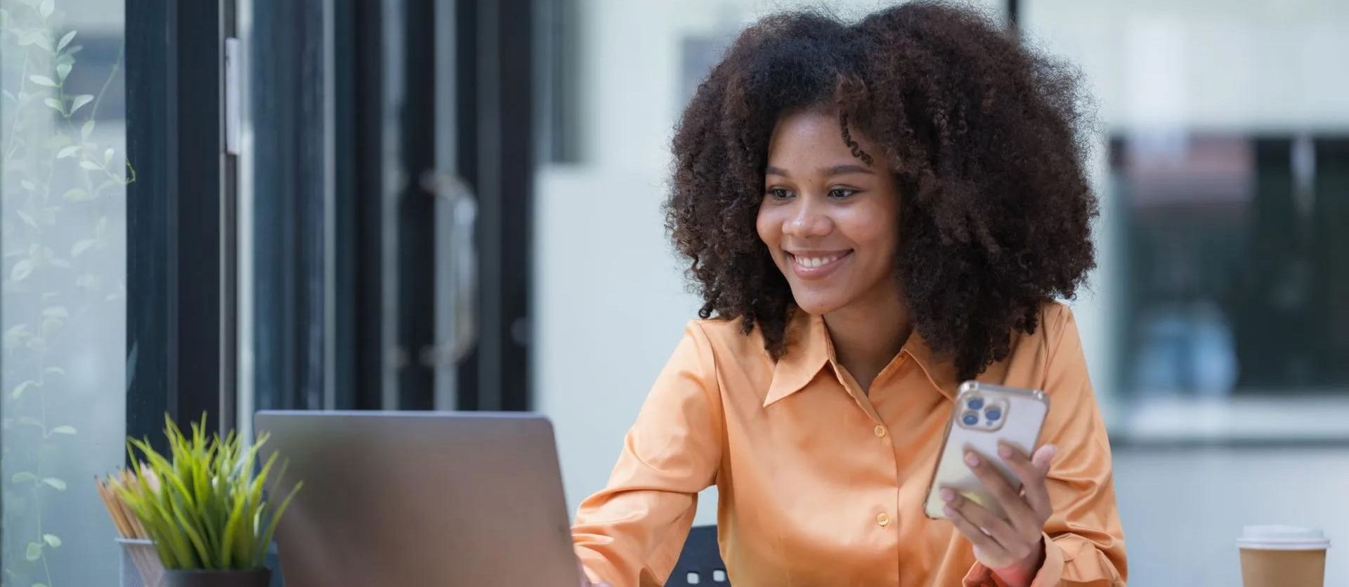 A woman is sitting at a desk using a laptop and a cell phone.