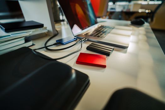 Un portátil y una libreta sobre un escritorio con cables, una cartera roja y un fondo de oficina borroso.