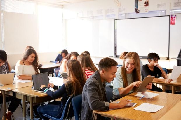 A group of students are sitting at desks in a classroom using laptops and tablets.