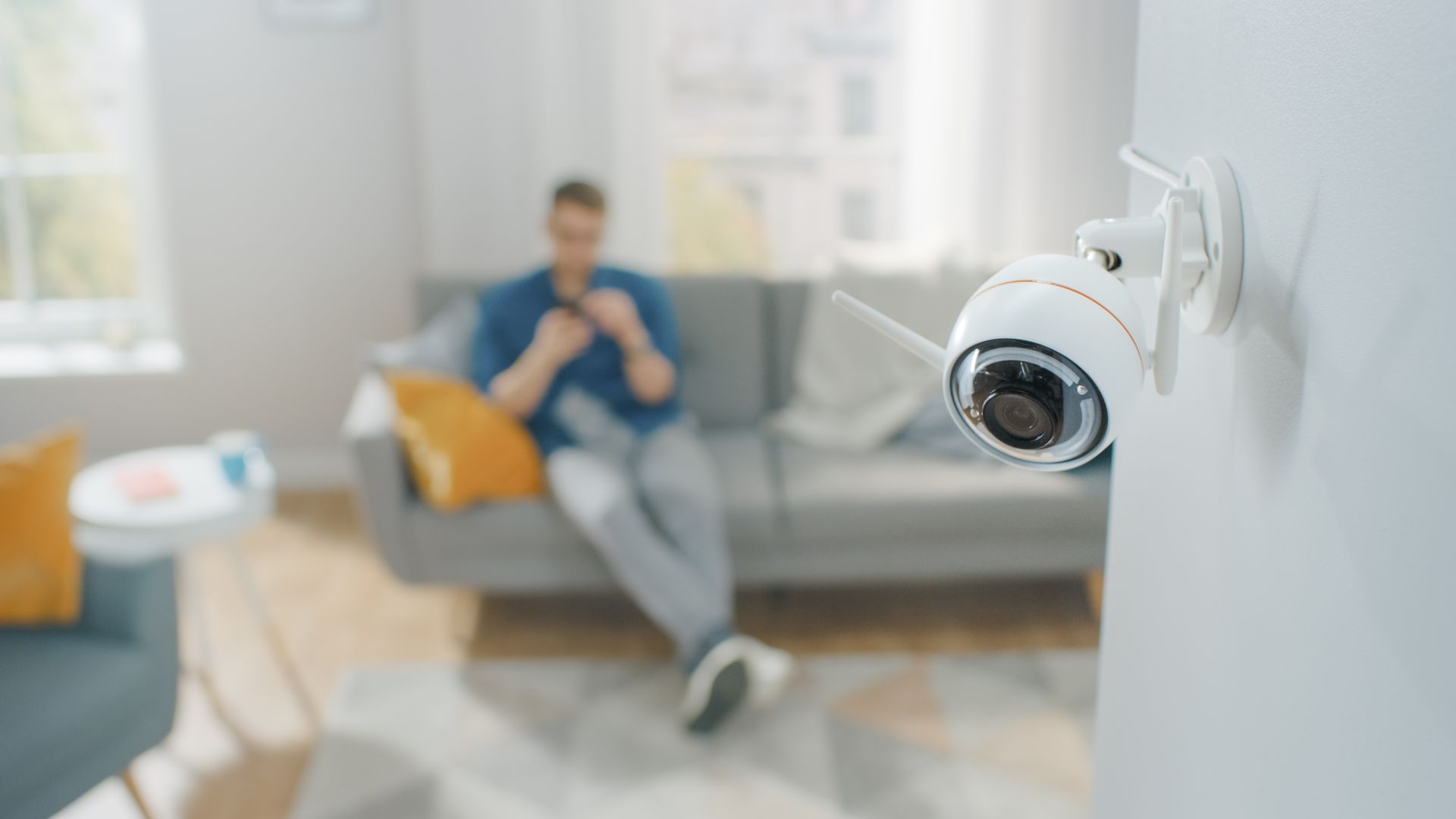 Security camera overlooking a living room with a person sitting on a sofa using a phone.