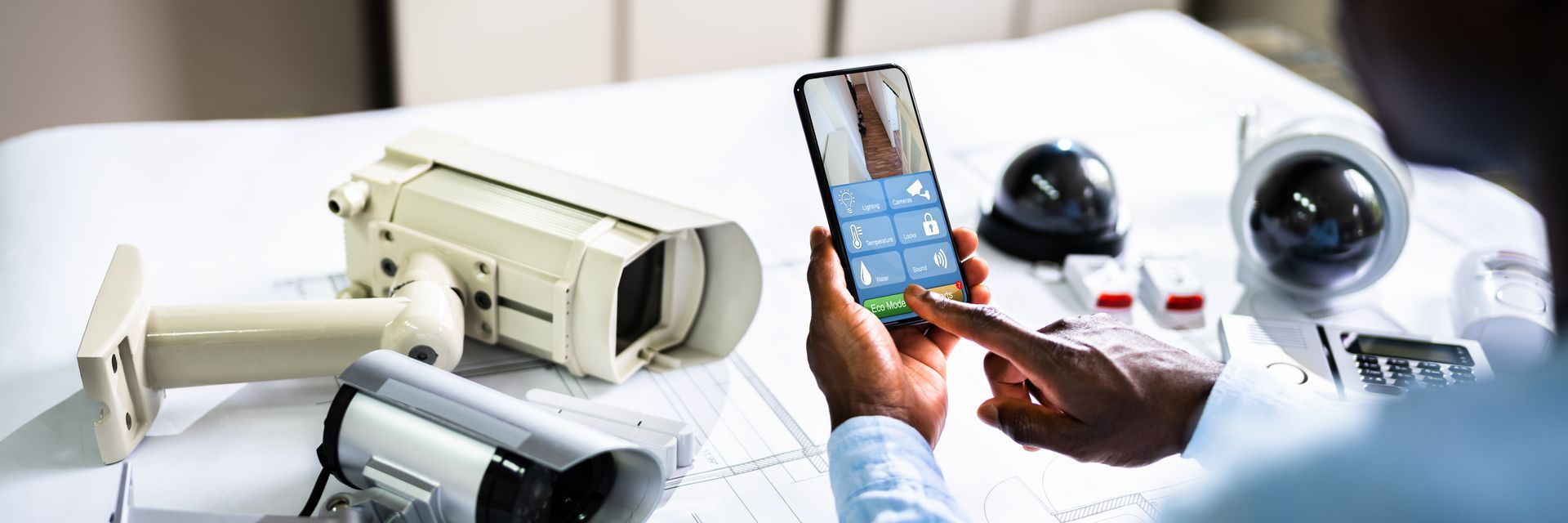 Hand holding a smartphone beside several security cameras on a white desk
