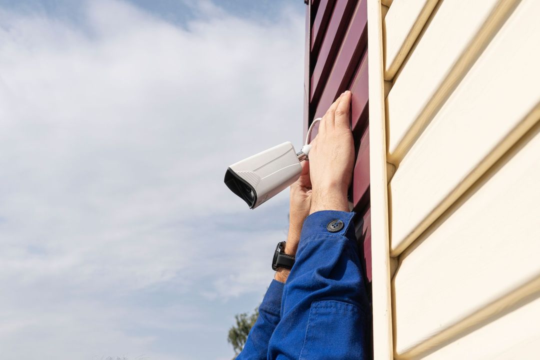 Hand holding a security camera near a house wall under a blue sky