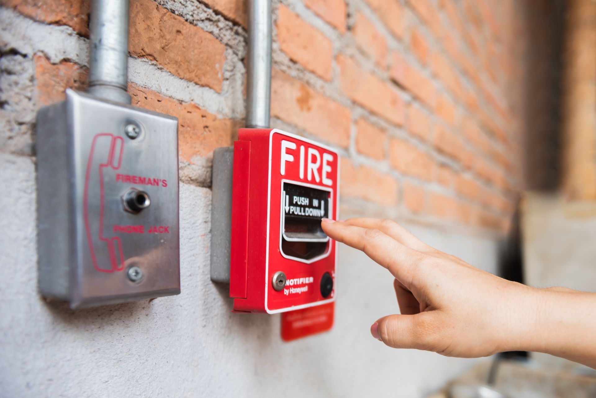 Hand pressing a red fire alarm pull station on a brick wall
