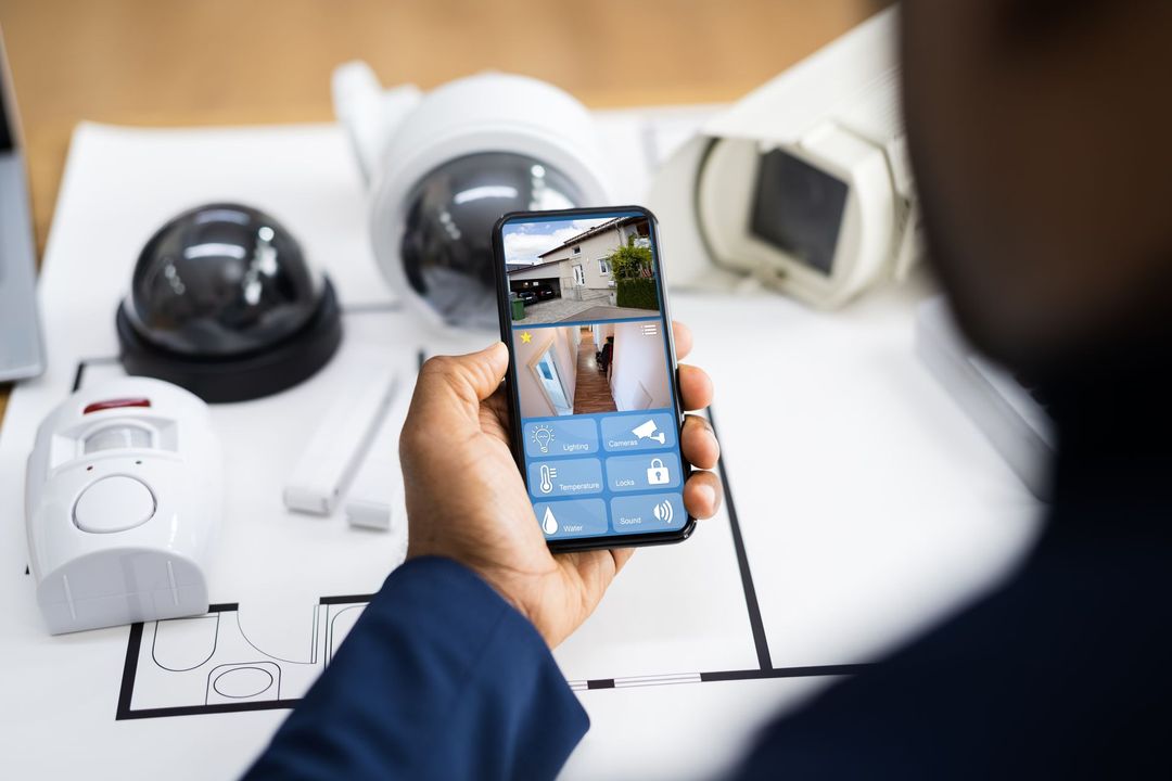 Hand holding a smartphone over a table with home security cameras and a monitor