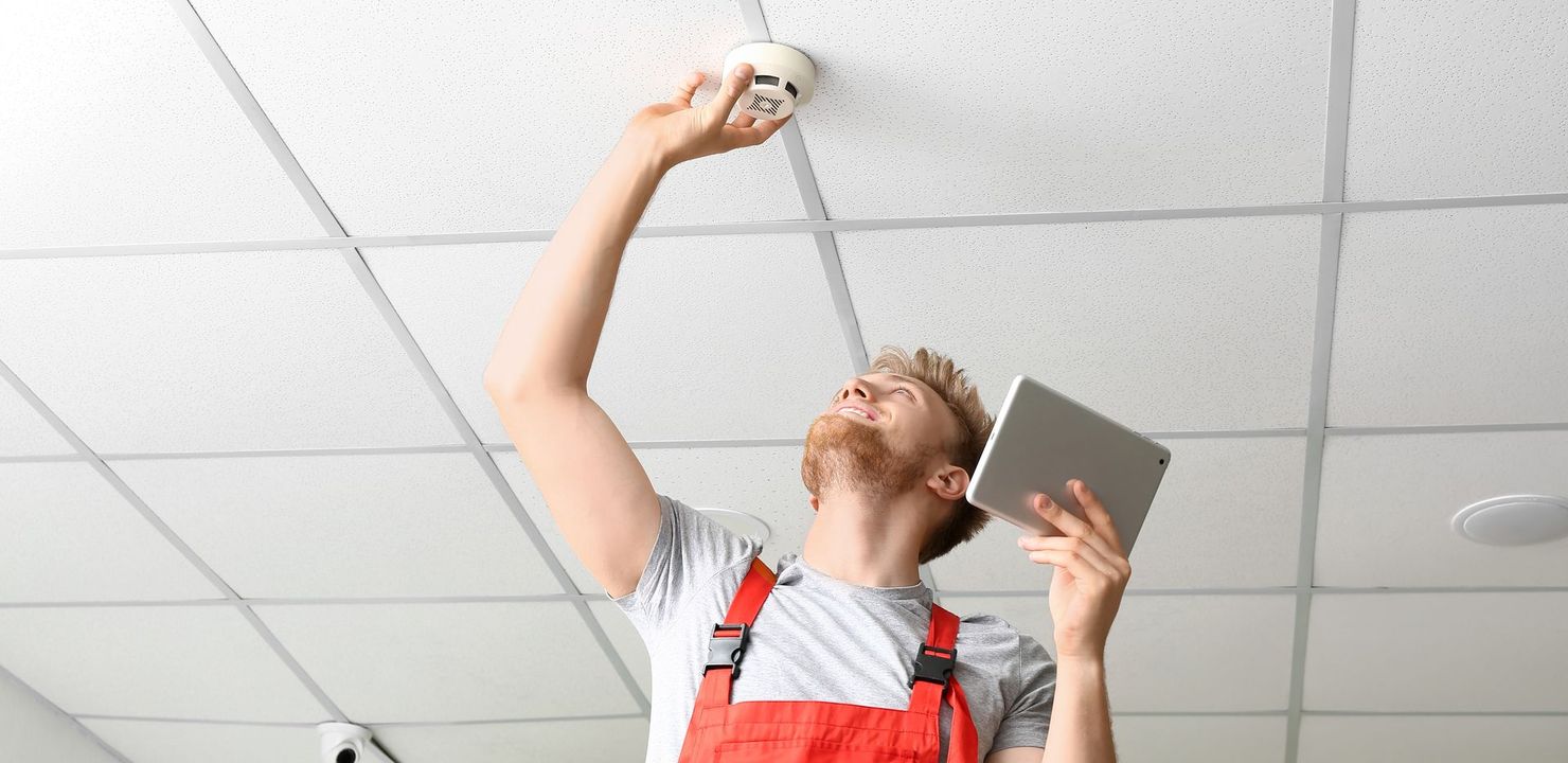 Worker in red overalls checking a ceiling smoke detector with a tablet