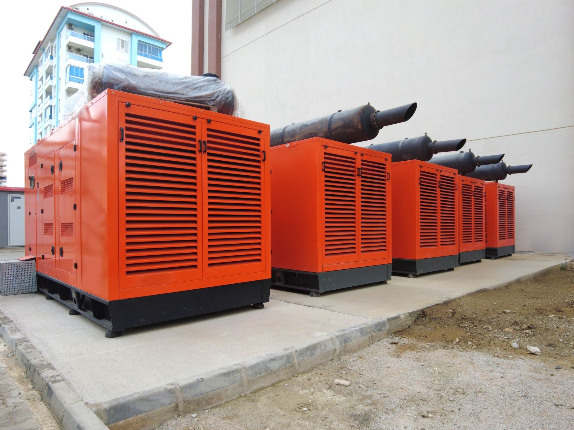 Row of orange industrial generators with black exhaust pipes outside a building