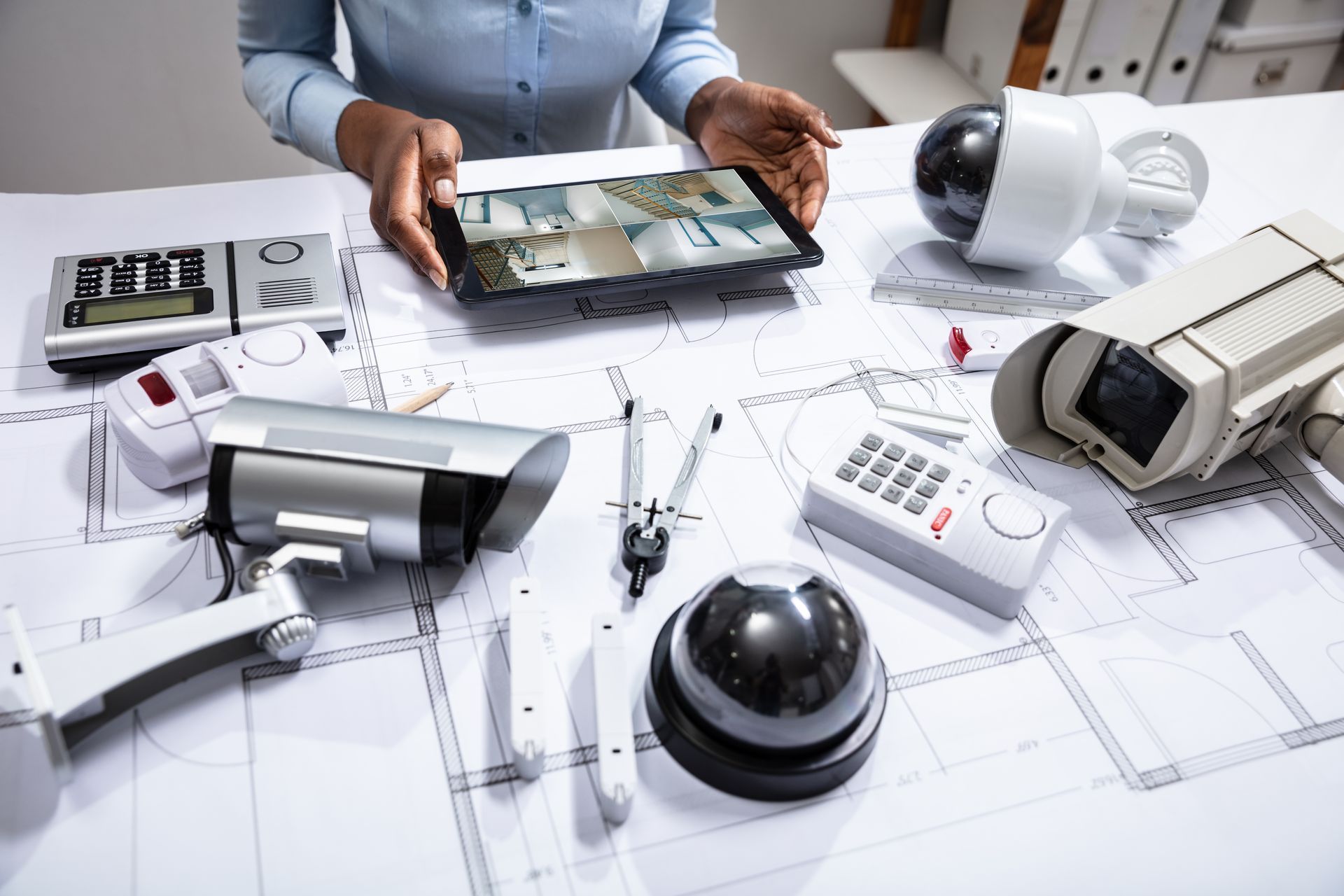 Surveillance equipment and blueprint plans on a desk, with a person holding a tablet showing a home security layout