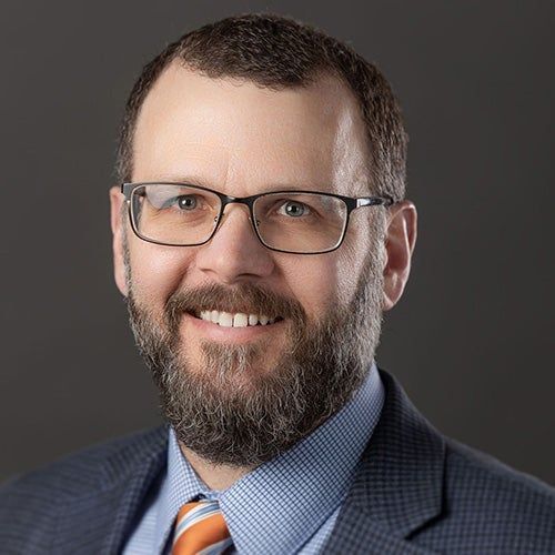 Head-and-shoulders portrait of a smiling man in glasses, suit jacket, blue shirt, and striped tie.