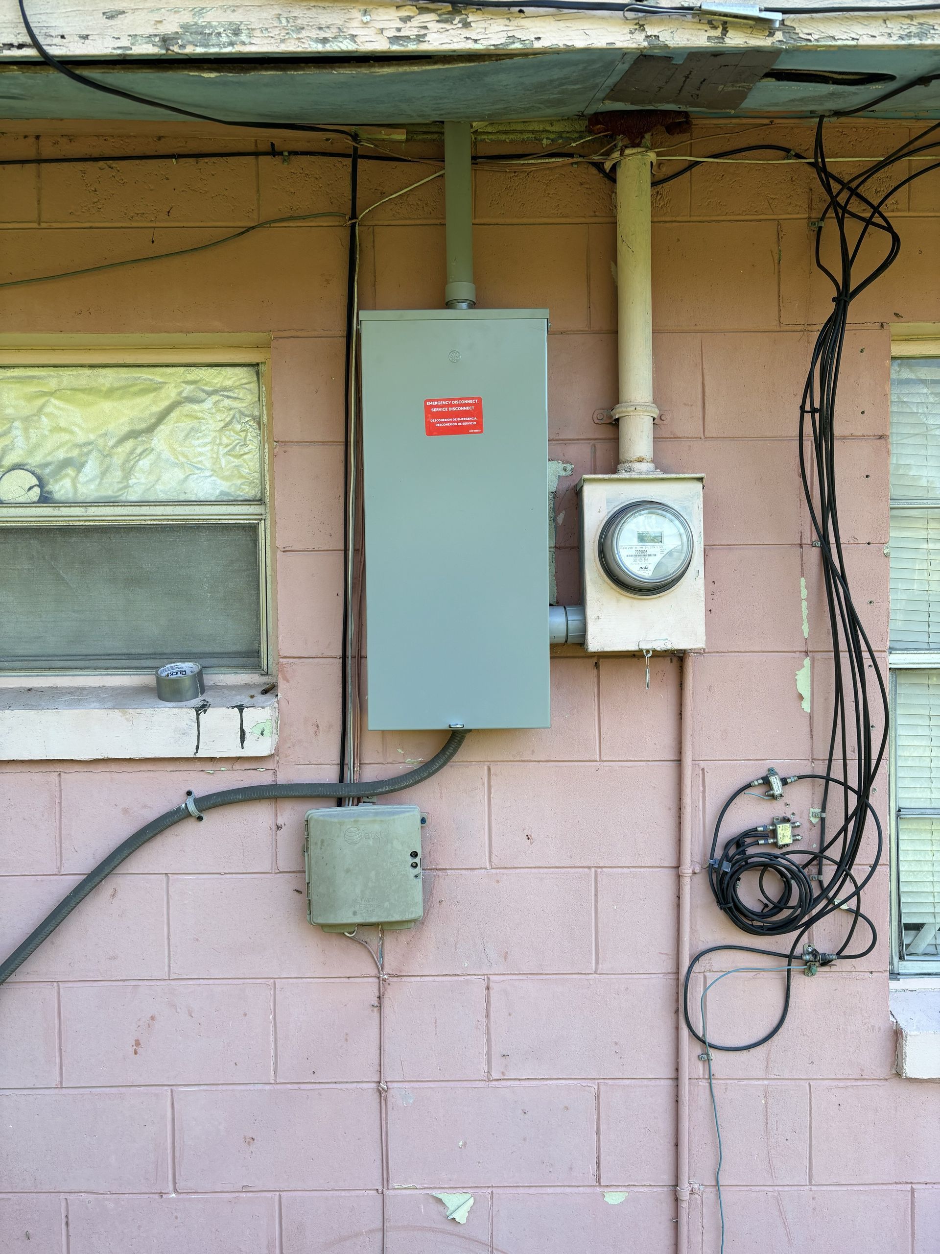 Exterior wall with electrical meter, gray panel, and hanging wires beside a window.