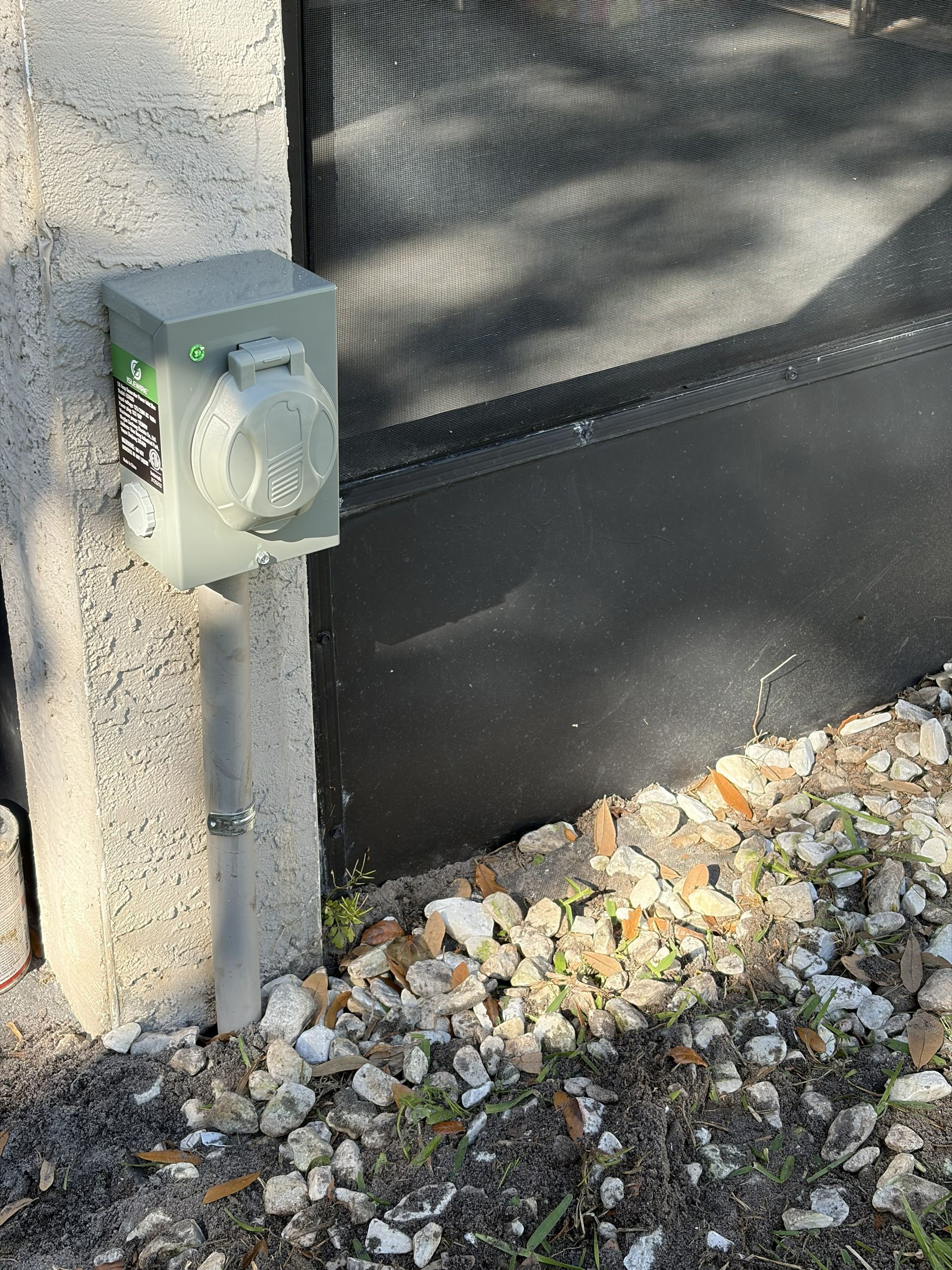 Small gray electrical box mounted on a concrete wall beside a black gate and gravel ground.