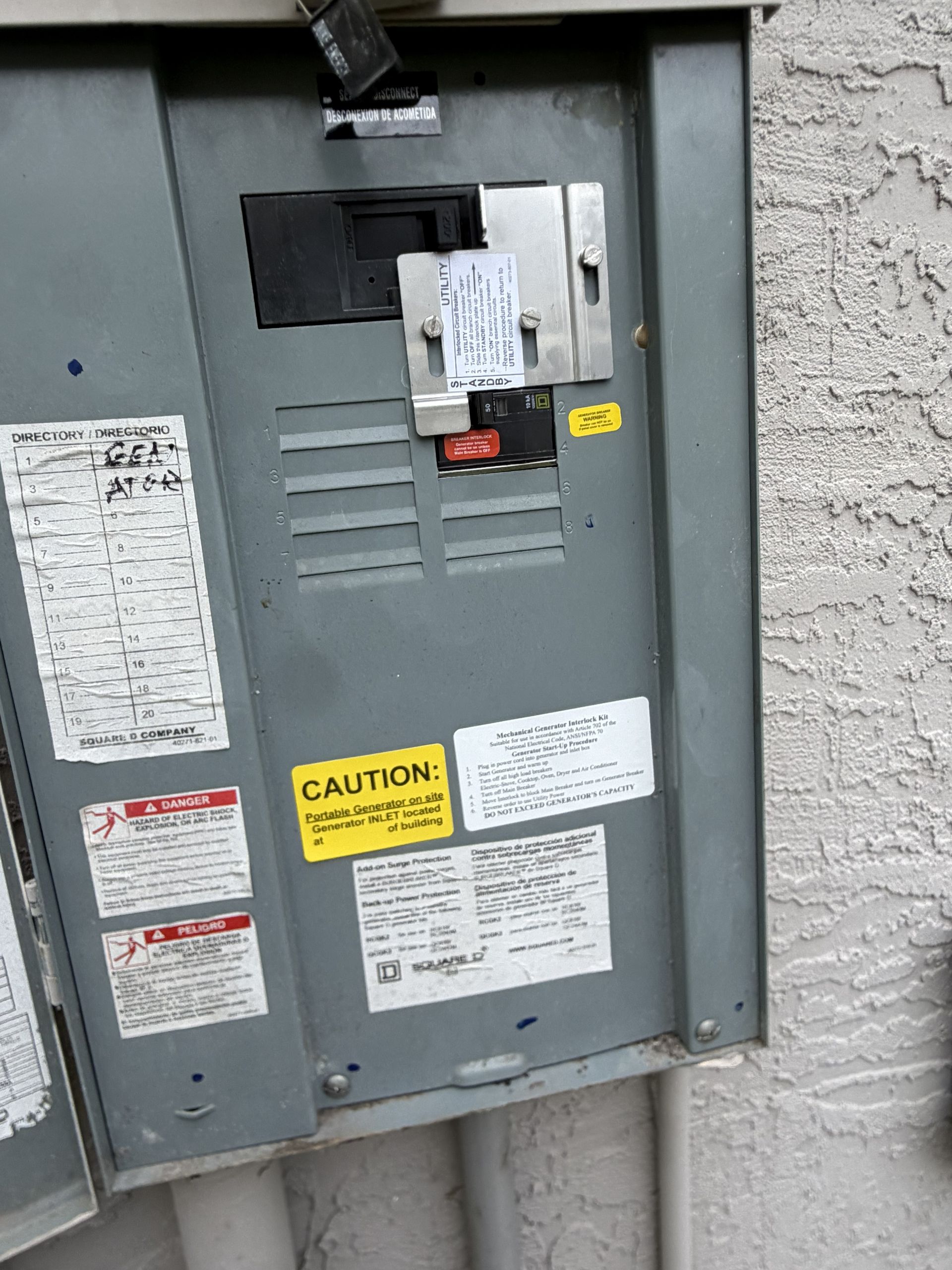 Gray electrical service panel with warning labels mounted on a stucco exterior wall.