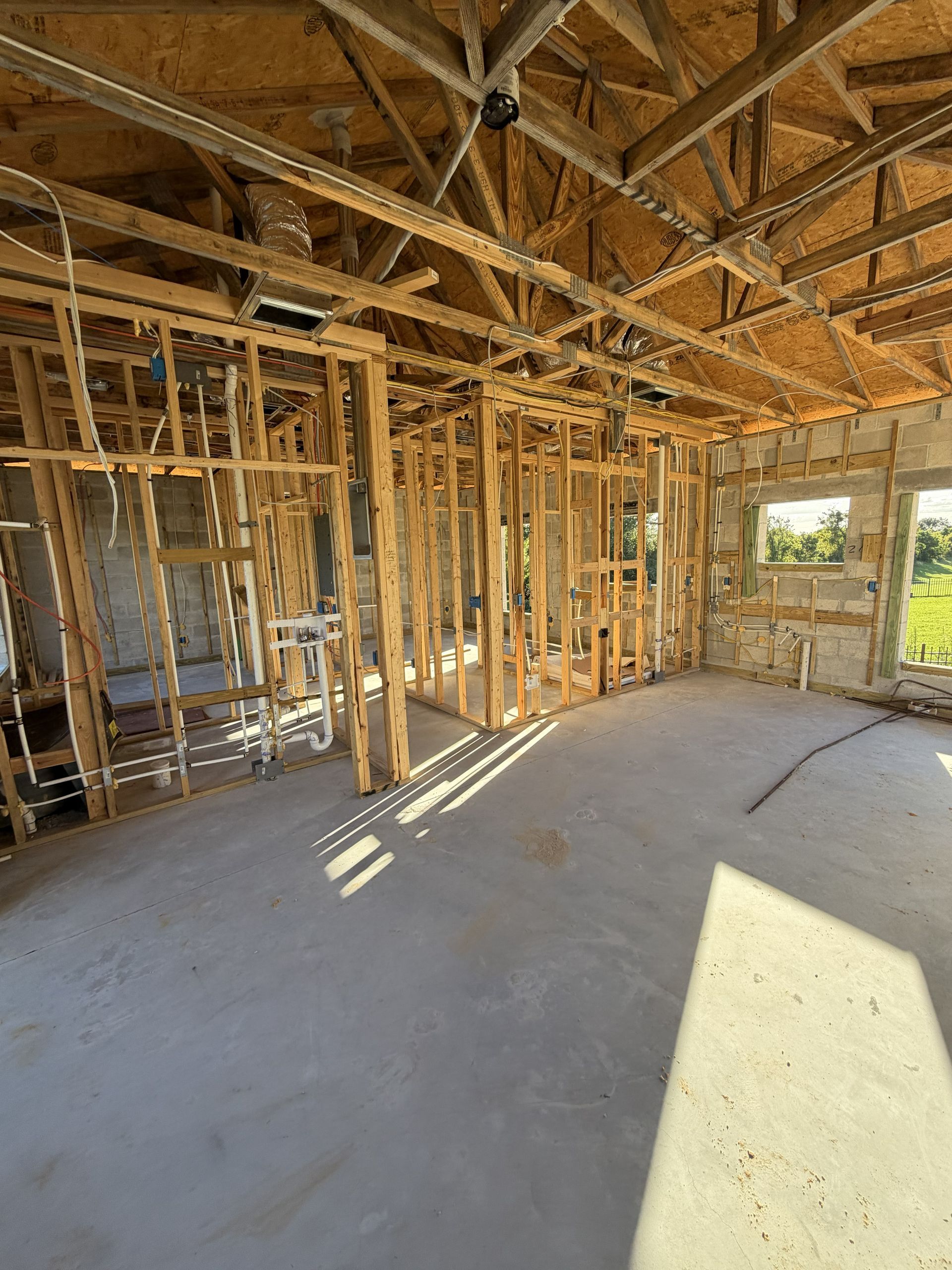 Unfinished house interior with exposed wooden framing, concrete floor, and sunlight streaming through openings