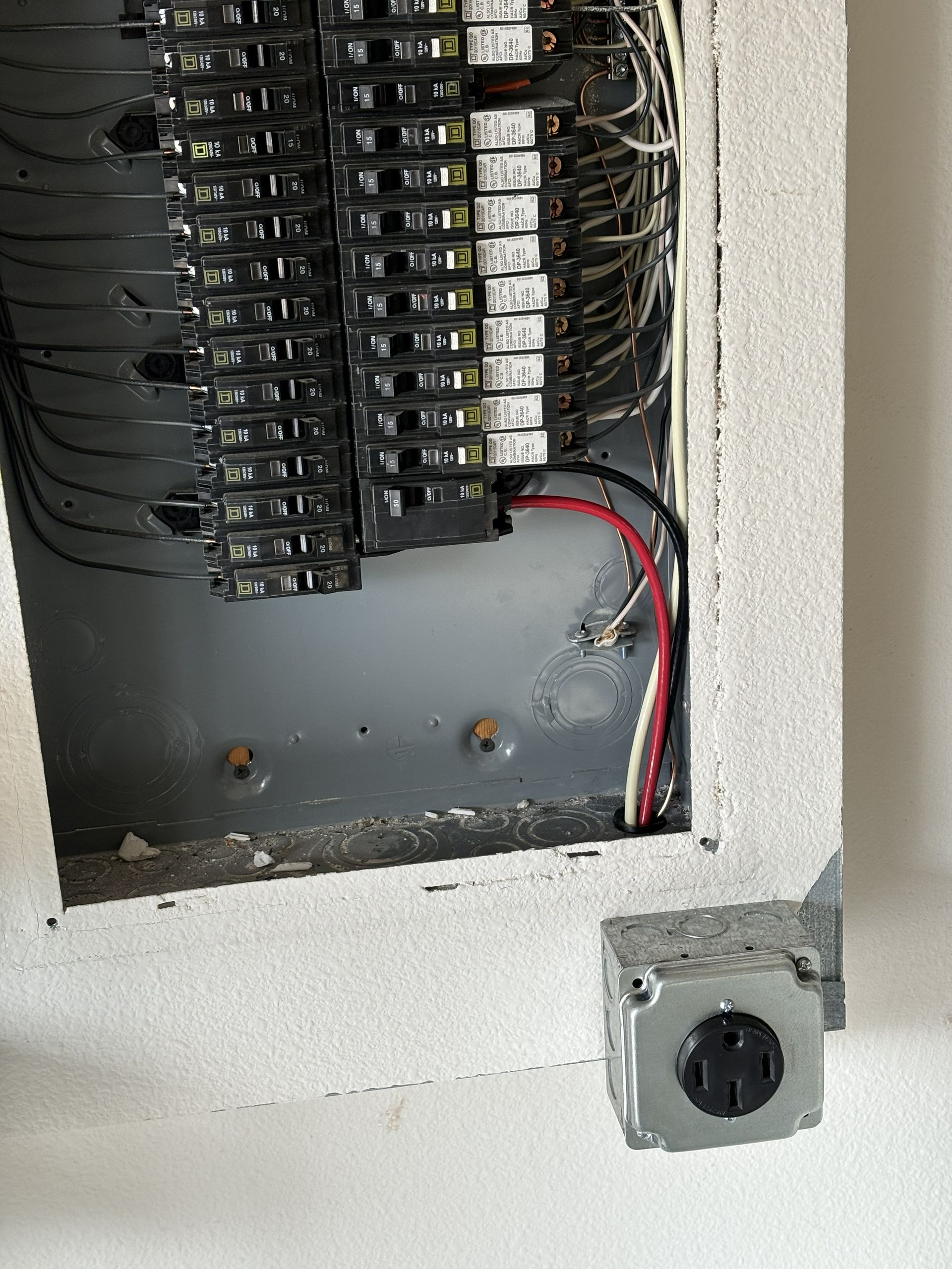 Open electrical breaker panel with red and black wires and a white wall outlet below
