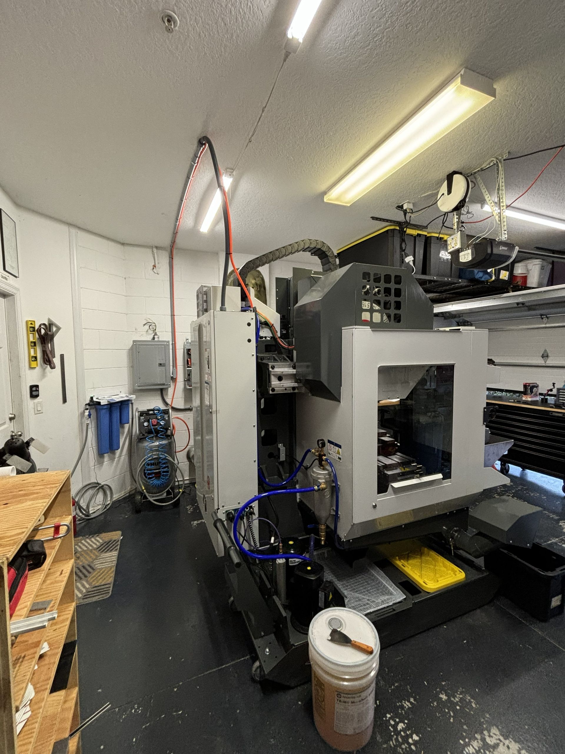 Industrial machine in a narrow workshop aisle, with tools, shelves, and overhead lights.