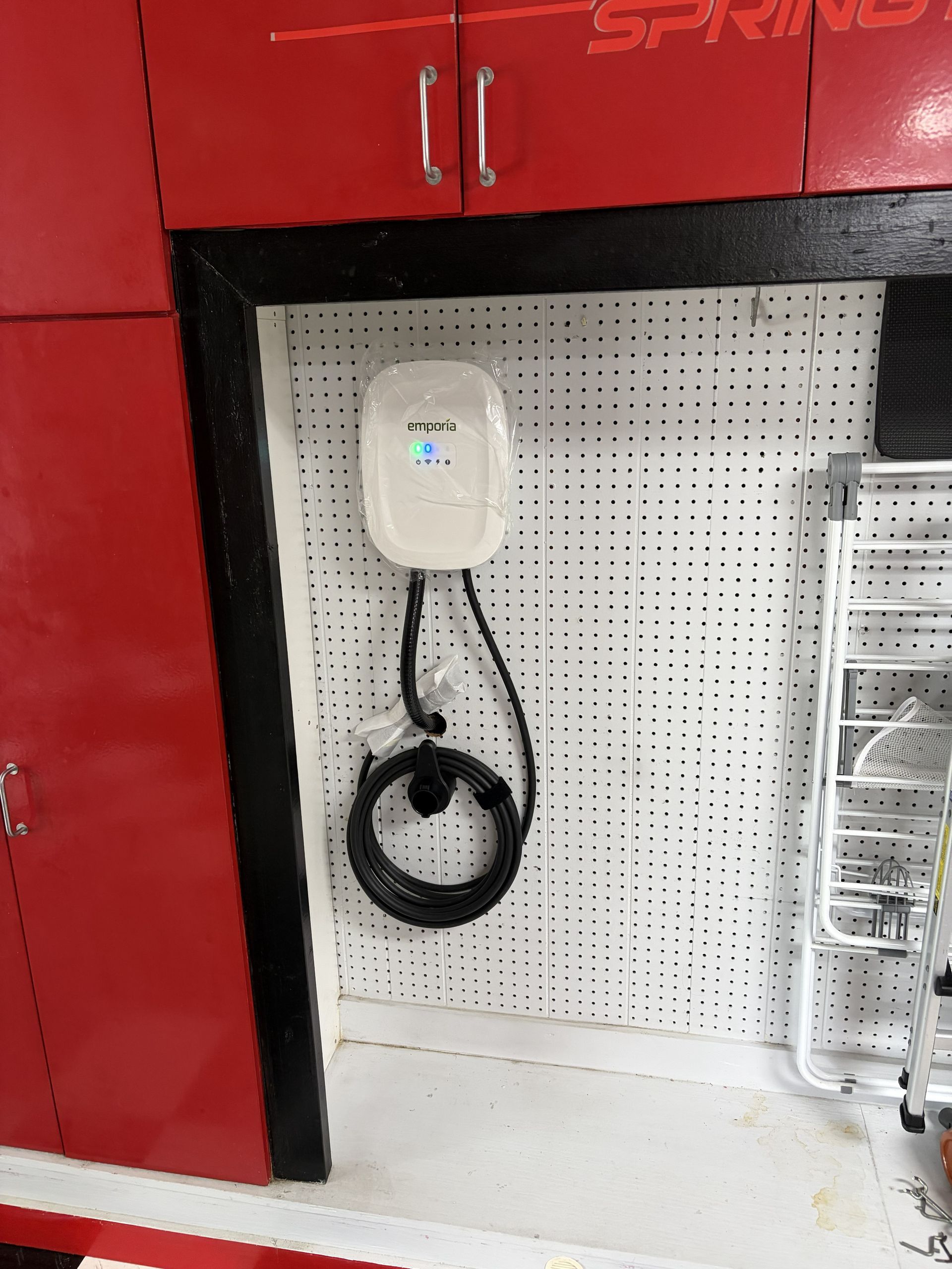 White wall-mounted charger with coiled black cable on a pegboard, between red cabinets in a garage.