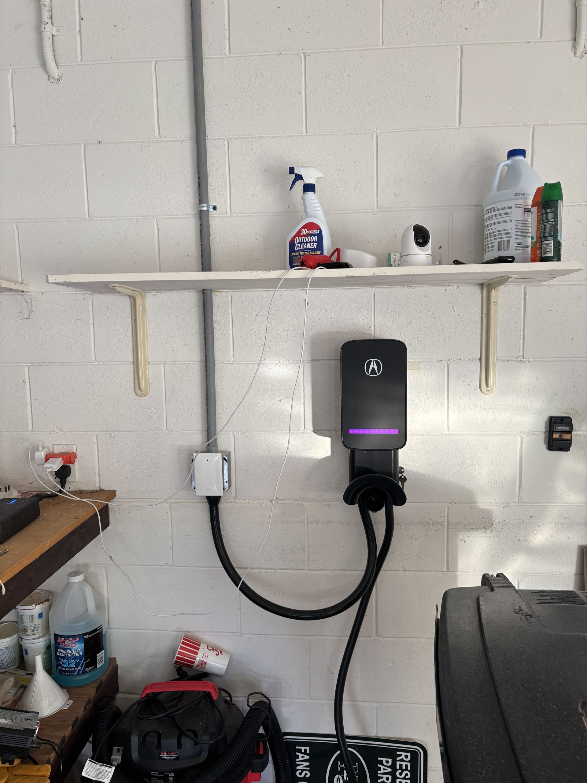 White brick wall with a shelf, cleaning supplies, a black wall-mounted device, and cables in a garage or workshop.