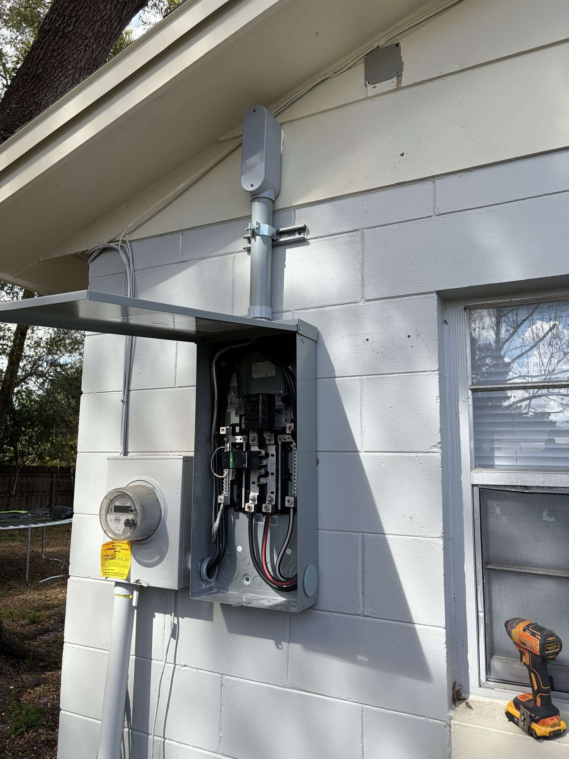 Open electrical panel on house exterior with exposed wiring, service meter, and drill on a ledge