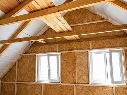 Interno di una soffitta non rifinita con travi in ​​legno a vista, pannelli isolanti e due piccole finestre.