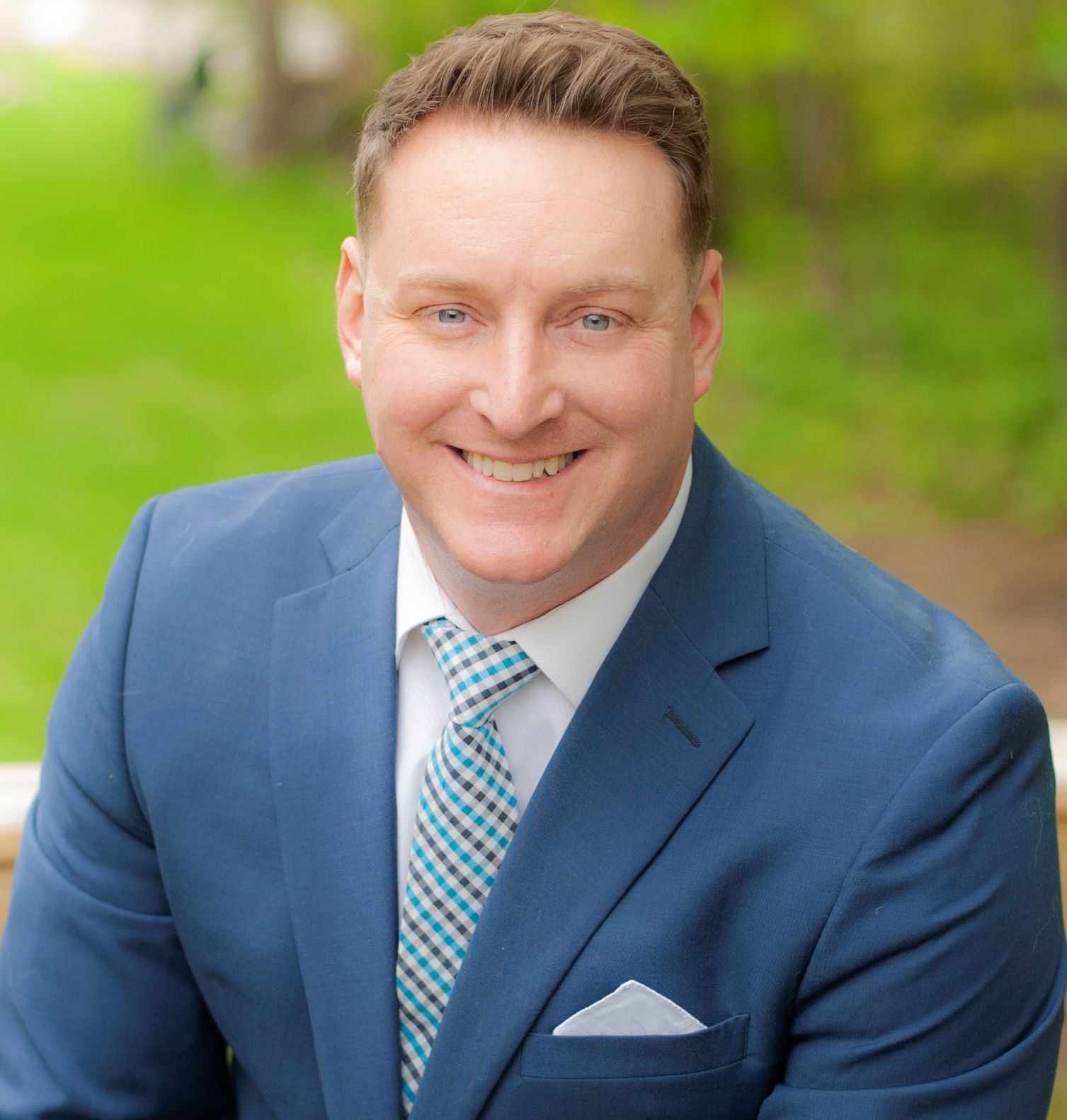 Man in blue suit, smiling, with a checkered tie, set outdoors with green background.