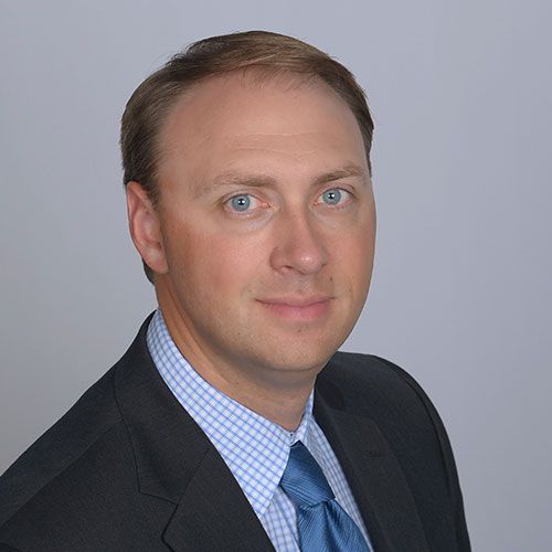 Man in a suit, light skin, blue eyes, smiling, headshot, against a gray background.