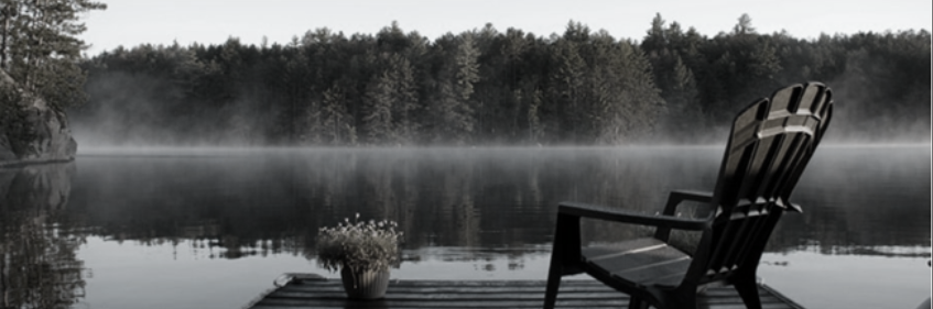 Black and white image of a lake with mist, a wooden dock, Adirondack chair, and trees.