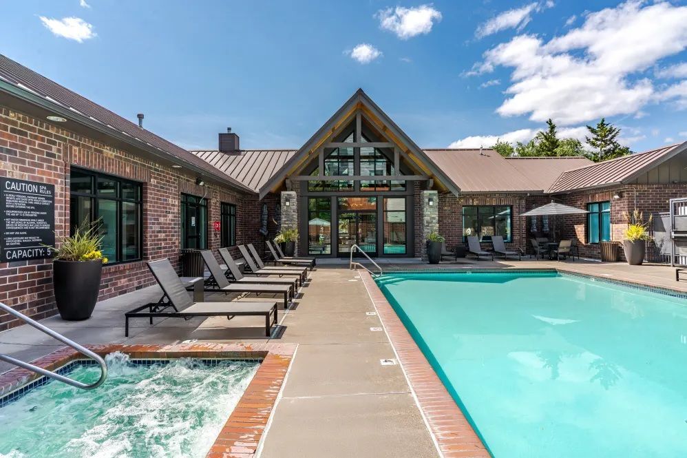 Poolside area with lounge chairs, hot tub, and building with large glass doors under a blue sky.