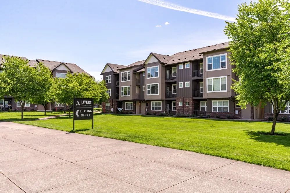 Apartment complex with multiple buildings, green lawn, and sign at The Avery at Orenco Station, offers apartments in Hillsboro, OR. Brown siding, blue sky.