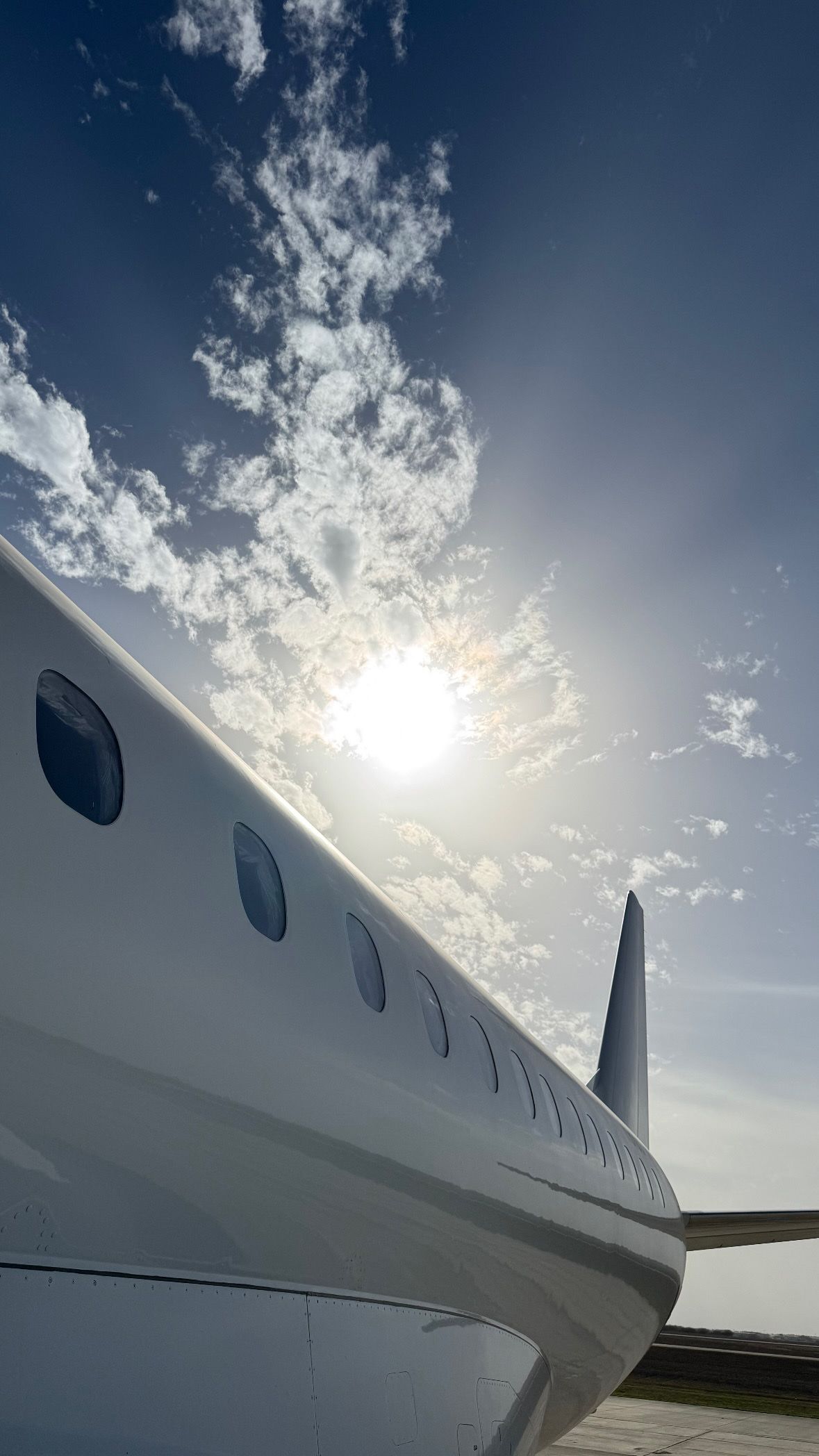 White airplane fuselage and wing under a bright sun and scattered clouds in a clear blue sky.