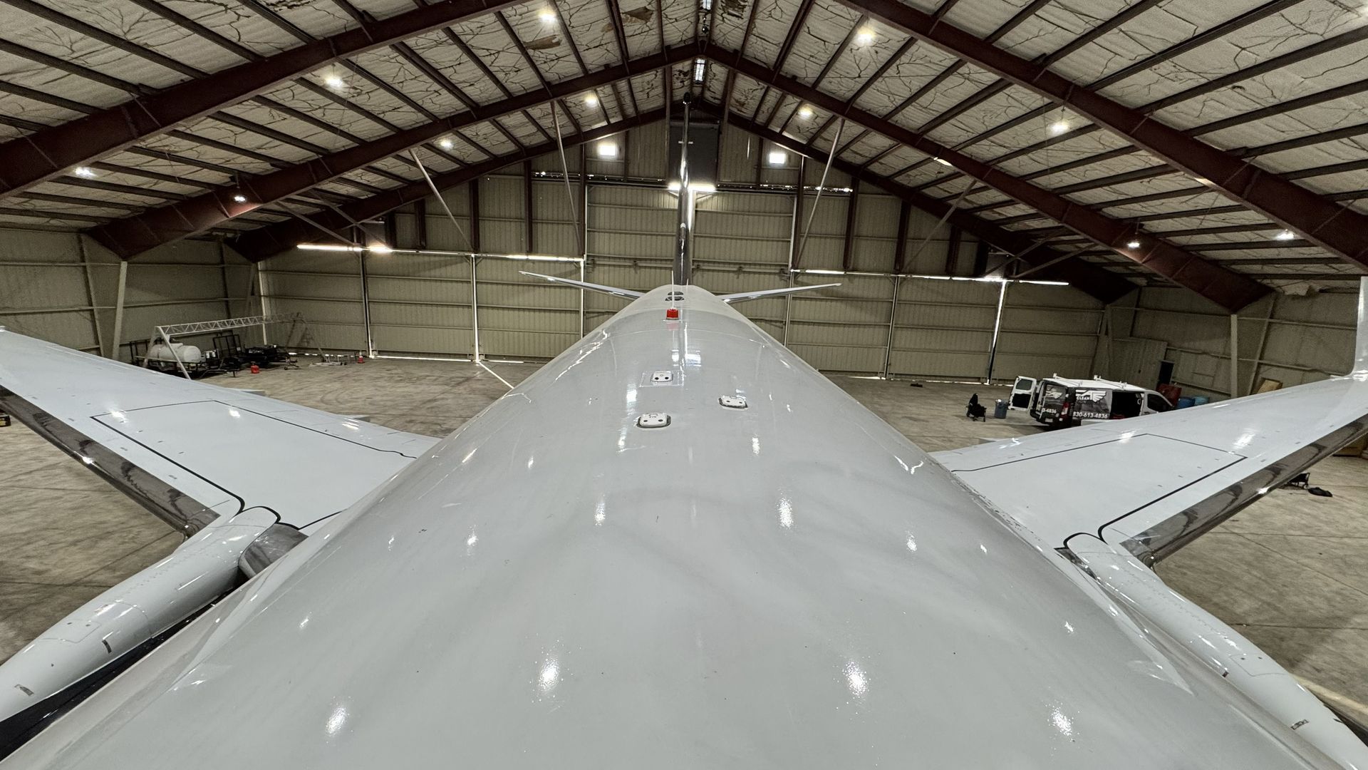 View from atop a white aircraft fuselage inside a hangar with open wings and roof trusses.