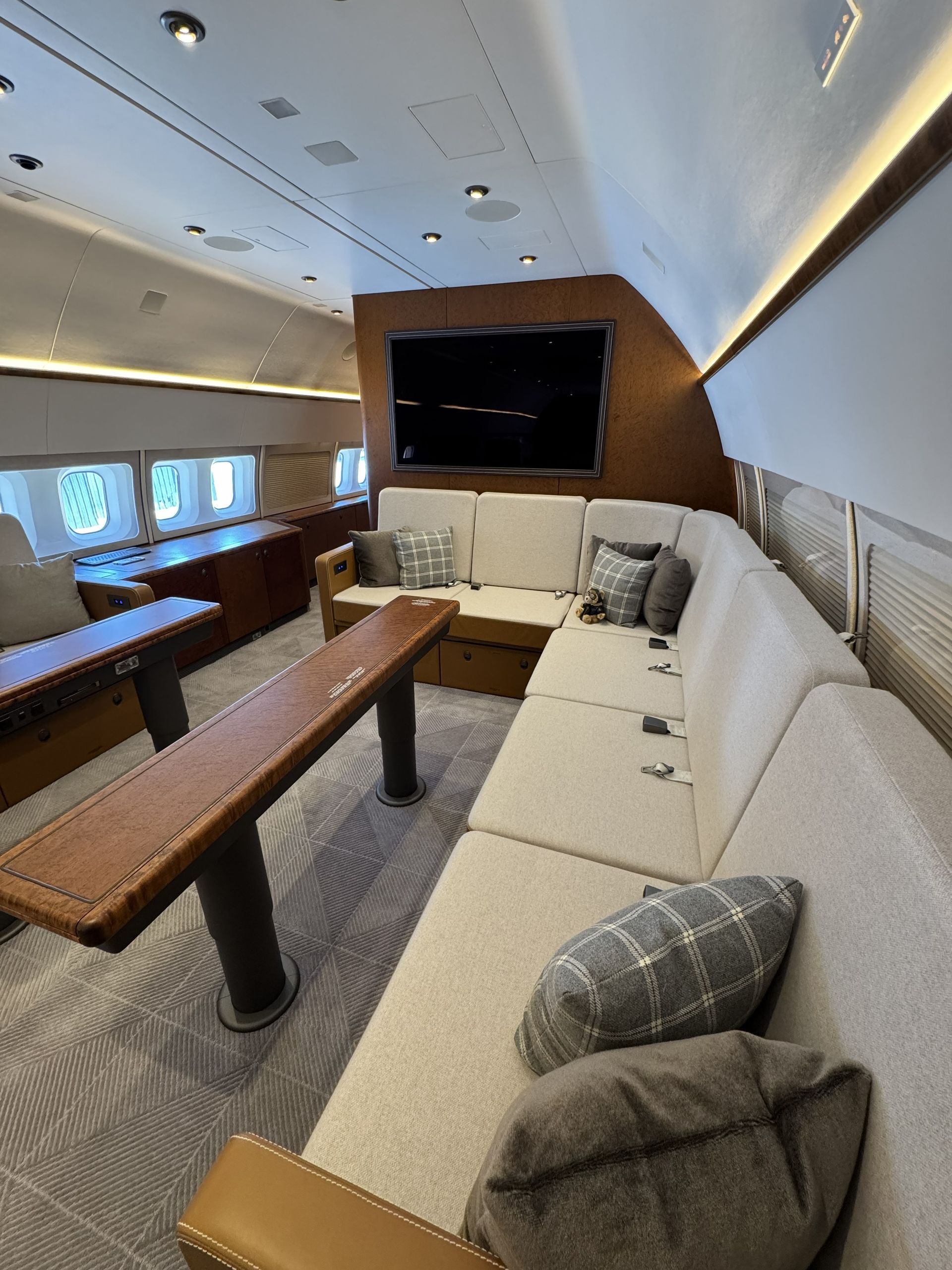 Airplane first-class suite with white seating, wood table, and large screen by windows.