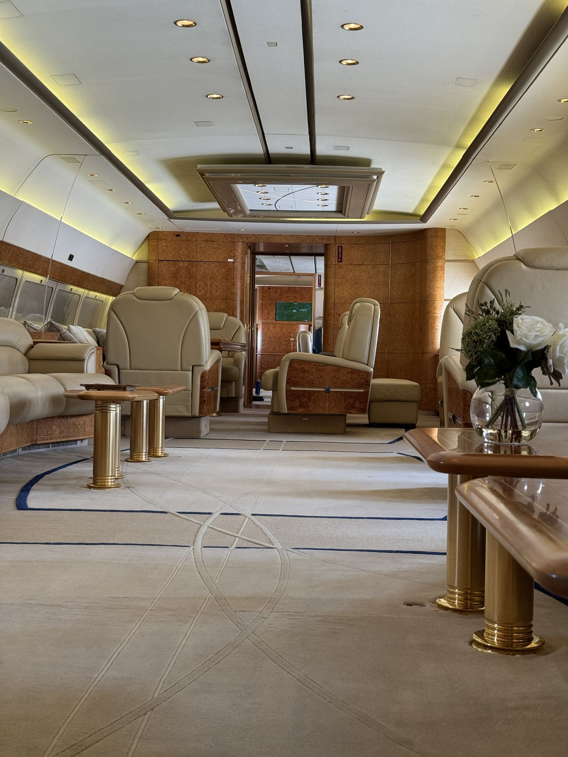 Luxury private jet cabin with beige seats, wood trim, carpeted aisle, and soft ceiling lighting